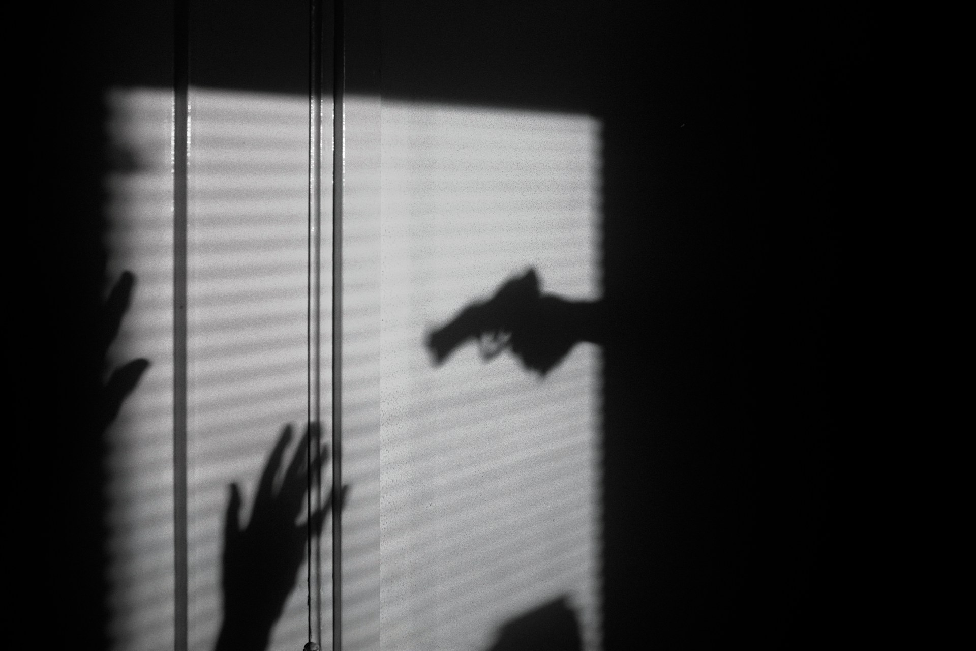 Foto dramática em preto e branco exibindo sombras projetadas em uma parede com persianas. Destaca-se a silhueta de uma mão empunhando uma arma de fogo apontada diretamente para a sombra de outras mãos erguidas em sinal de rendição ou defesa. A cena tensa simboliza a violência armada e ilustra um artigo jurídico sobre a nova legislação que endurece as regras para criminosos que cometem homicídio qualificado contra agentes de segurança pública, Forças Armadas ou seus familiares.